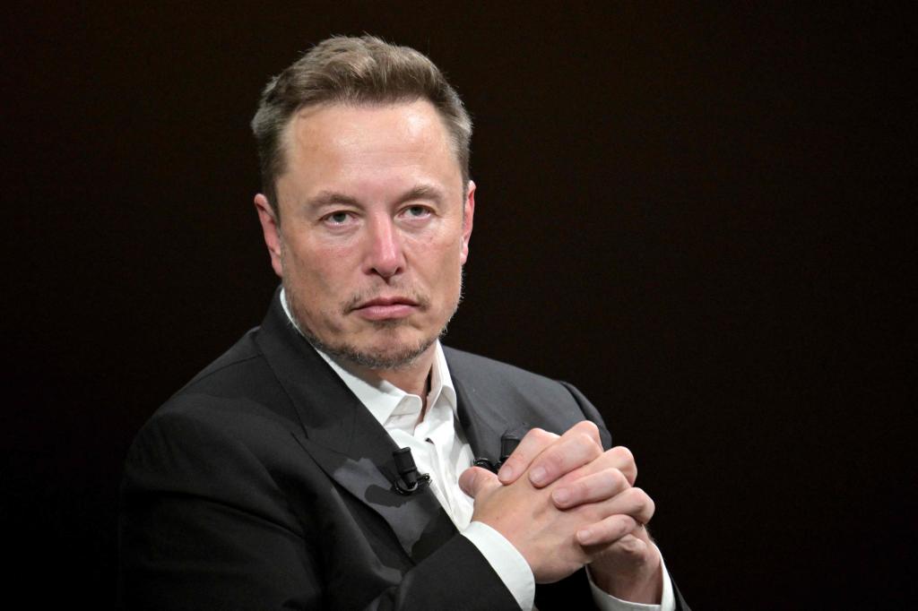 Elon Musk, in a suit, stands looking at the camera with his hands clasped together.