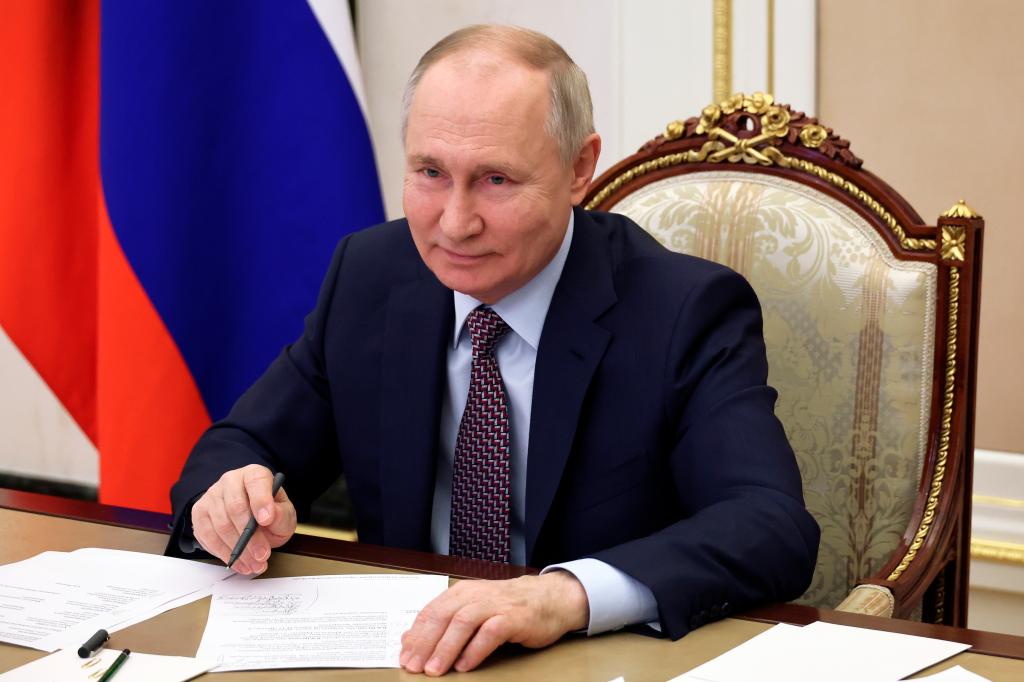 Russian President Vladmir Putin sits smiling at a desk with papers in front of him and a pen in his right hand.