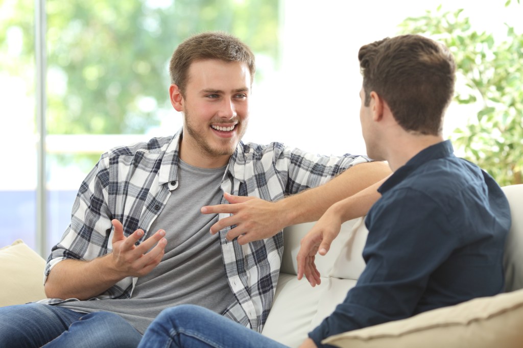 Two friends talking sitting in a couch in the living room with a window in the background at home