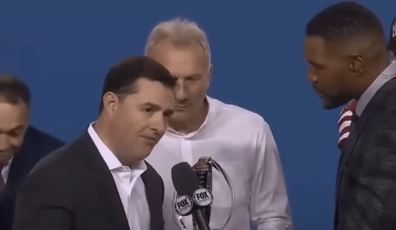 Joe Montana stands with the trophy while Michael Strahan (r) interviews Jed York (l).