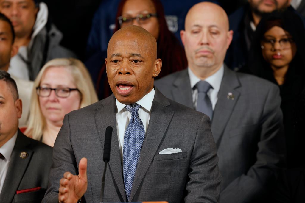 Mayor Eric Adams standing at press conference speaking with microphones and NYC officials around him including a female police officer in uniform.