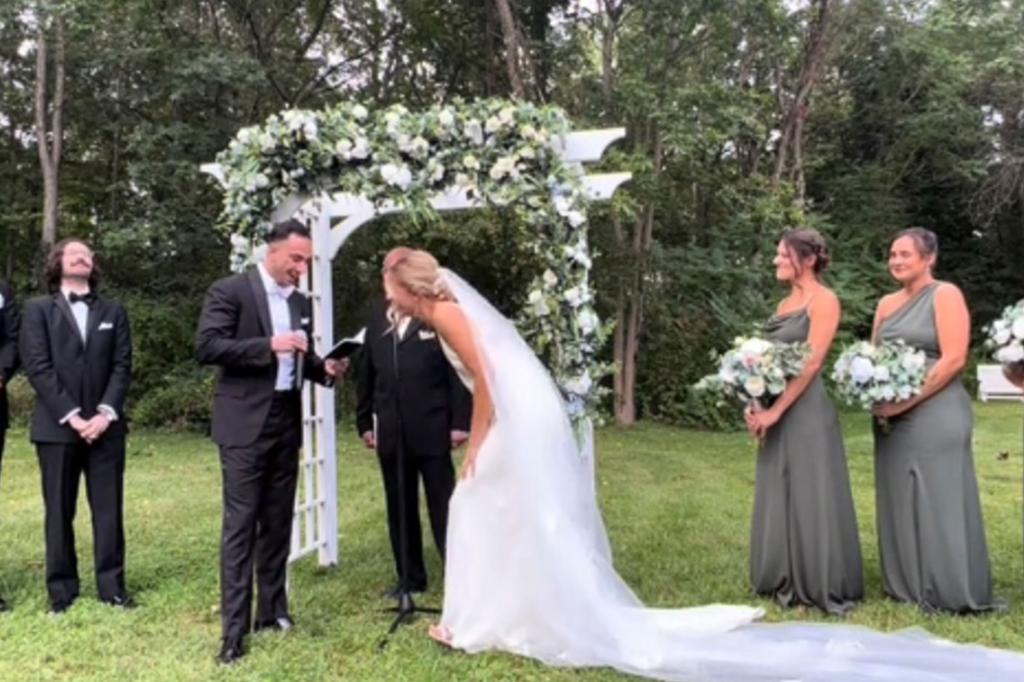 Bride bends over laughing as groom holds vows during outdoor ceremony. 