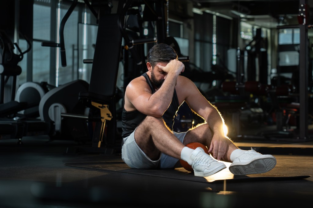 a tired man sitting on a mat after overtraining at a dark gym with other fitness enthusiasts in the background