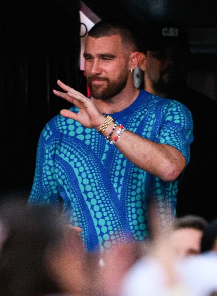 Travis Kelce, a man in a blue shirt, standing on a stage with fans in the background.