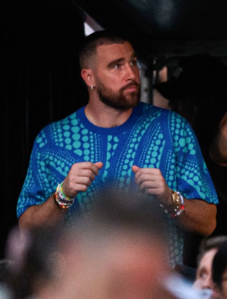 Travis Kelce, a man in a blue shirt, standing on a stage with fans in the background.