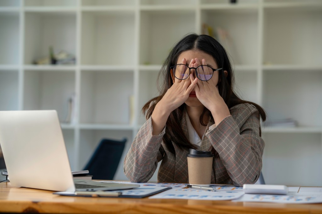 a tired woman covering her eyes with her hands sitting at her desk, experiencing office syndrome