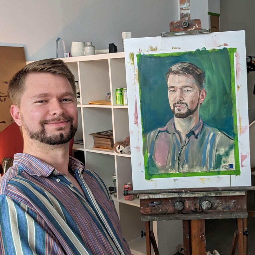 Owen MacDonnell standing beside his portrait, painted by Brooklyn artist Rusty Zimmerman, at a street-level gallery in Industry City