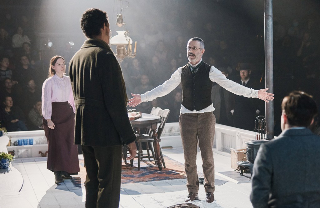 Actors Jeremy Strong and Victoria Pedretti in a scene from the play 'An Enemy of the People', with a man standing on a platform, arms outstretched. Photo by Emilio Madrid.