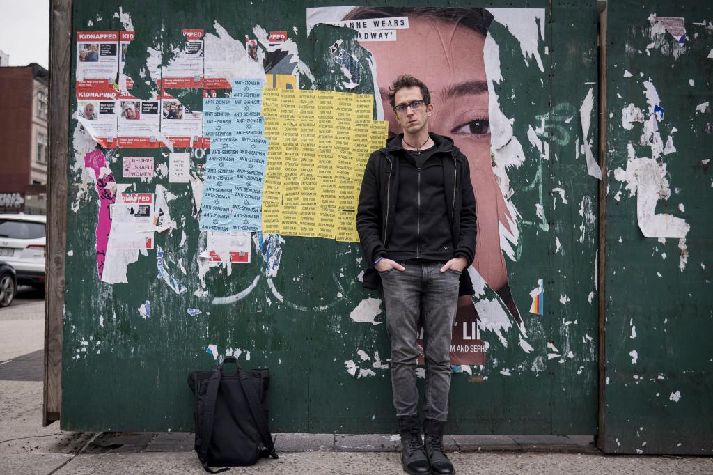 Columbia University researcher Shai Davidai leans against a green plywood wall covered in ripped ads