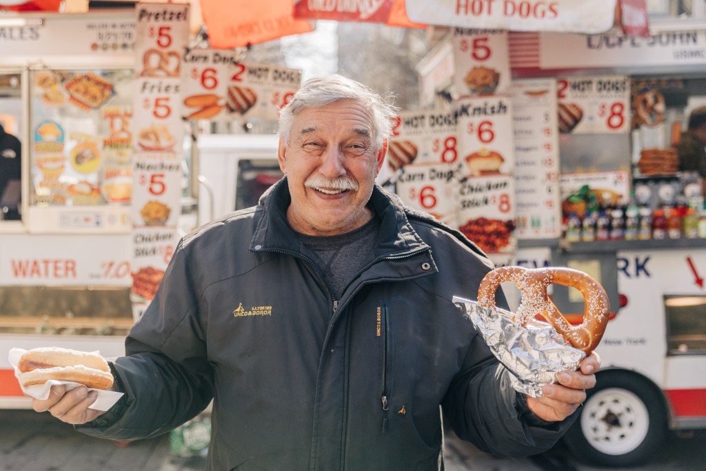 Dan Rossi, the NY Hot Dog King, holding a pretzel and hot dog outside his cart. 