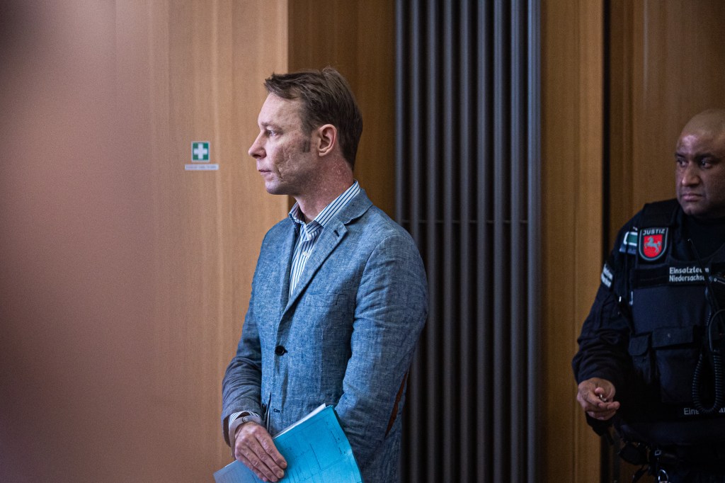 Christian Brueckner appears in court wearing a light blue sport jacket and stripped shirt, carrying a blue folder.  A uniformed officer is behind him.