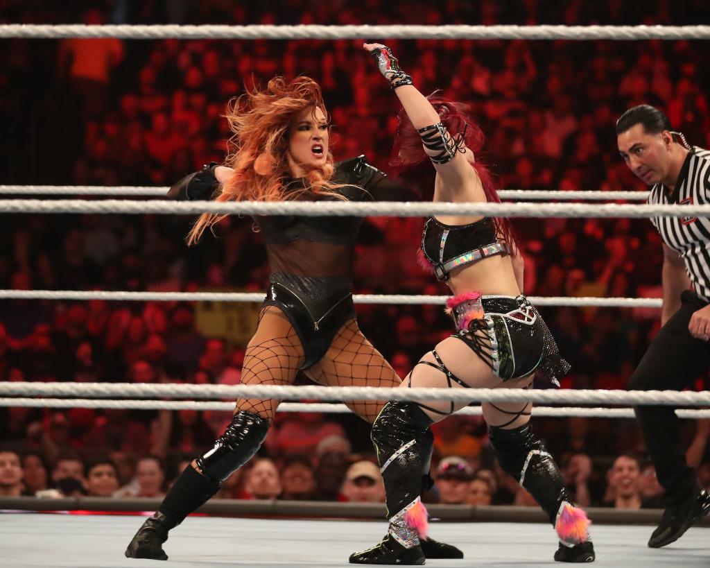 becky lynch wrestling in a black leather and sheer panel body suit with black knee high boots and her red hair flying while a referee looks on