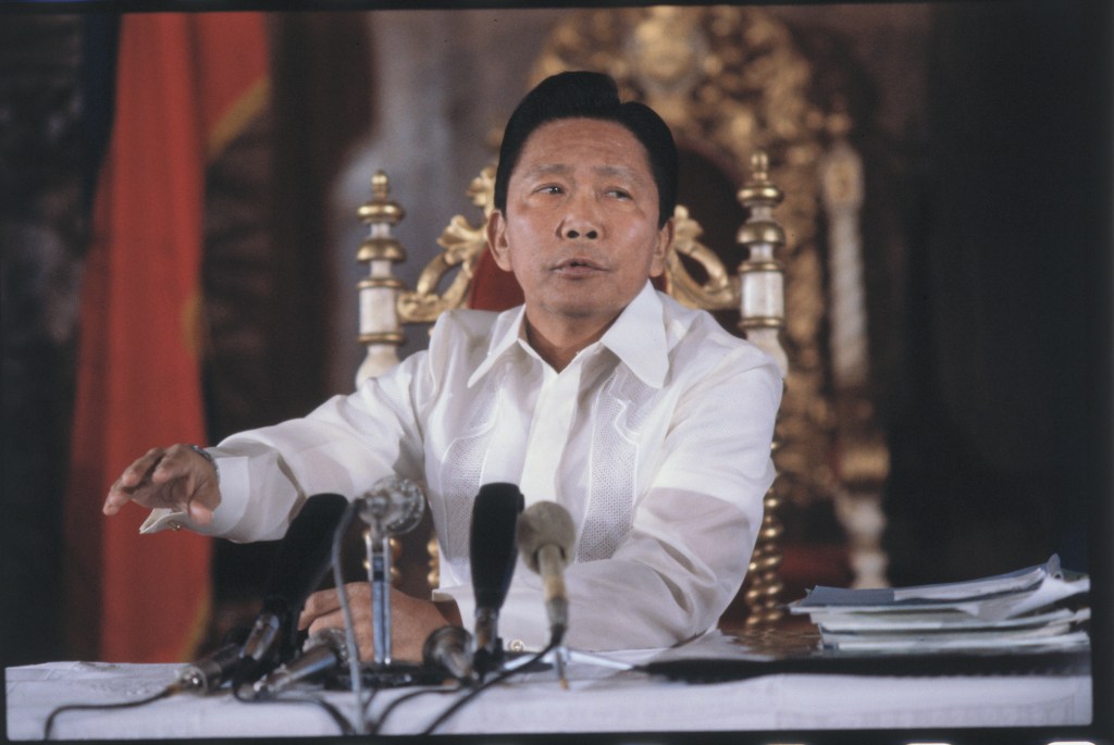 Ferdinand Marcos in 1980. He sits at a desk with several microphones in front of him. 