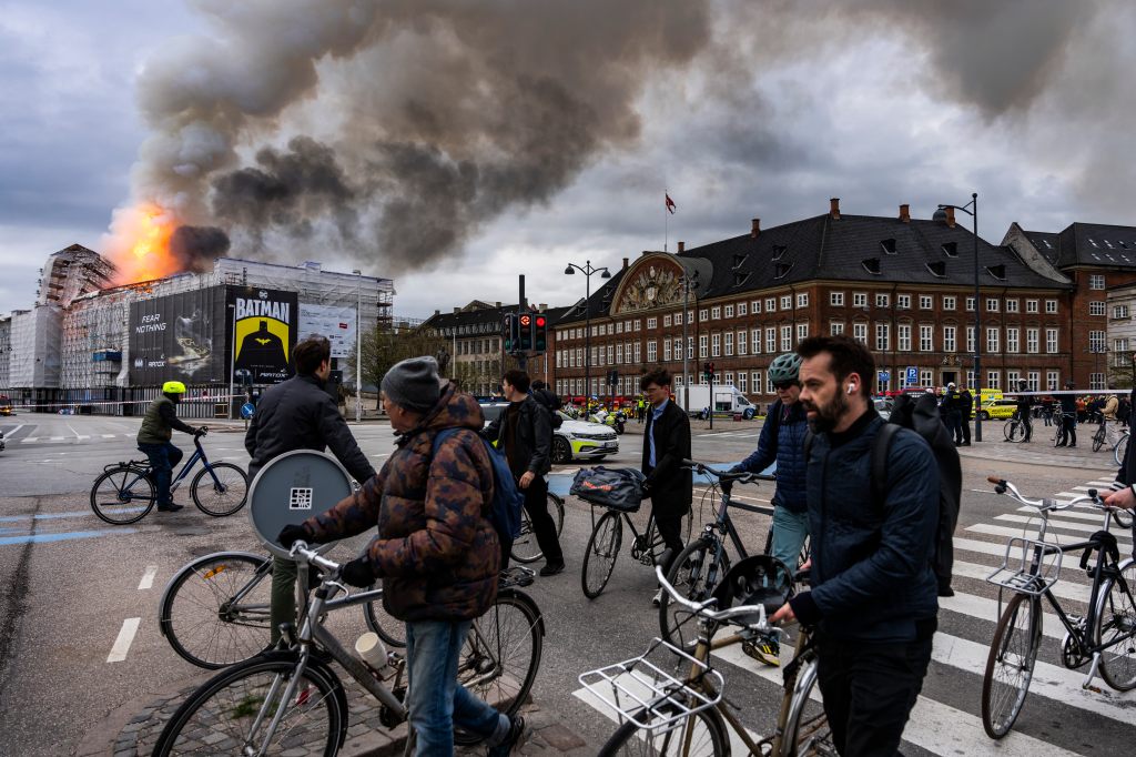 People riding bicycles in Copenhagen with smoke rising from the Old Stock Exchange building on fire in the background