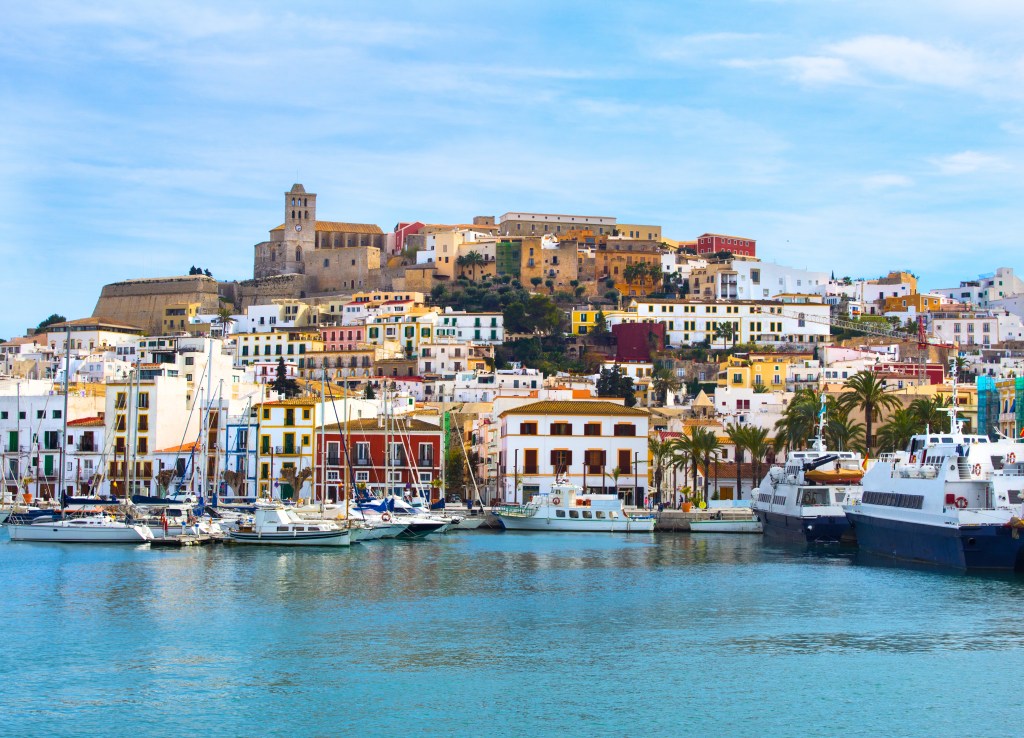 The island of Ibiza, crowded with buildings built into the hillside and yachts in the harbor, seen from the water.