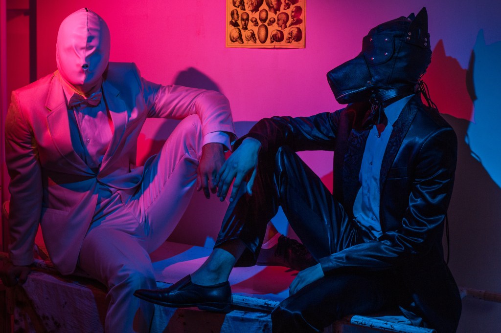 two guys, one in a black suit and a dog mask, one in a white suit with a white latex mask, is sitting on a stretcher