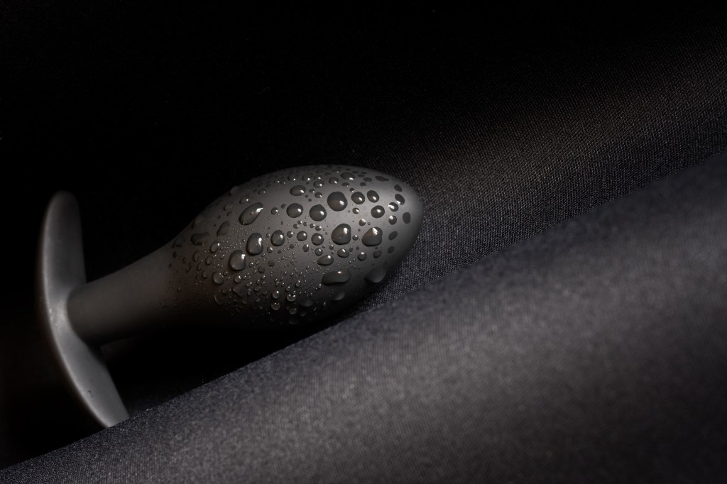 A wet black butt plug lies on a dark surface. Moisture or lubricant drops. Sex toy and pleasure. Diagonal