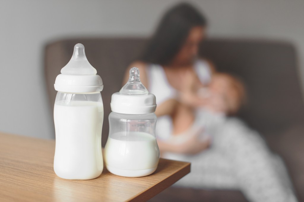 Bottles filled with breast milk on a table, with blurred background of a mother lovingly breastfeeding her baby