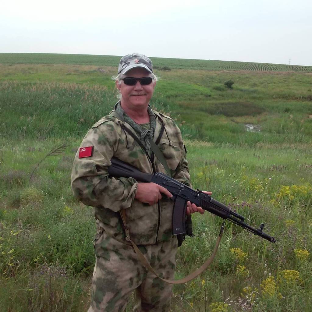 American Russell Bentley, dressed in a camouflage uniform and sunglasses, smiles while posing in a field in Ukraine, holding a long gun.