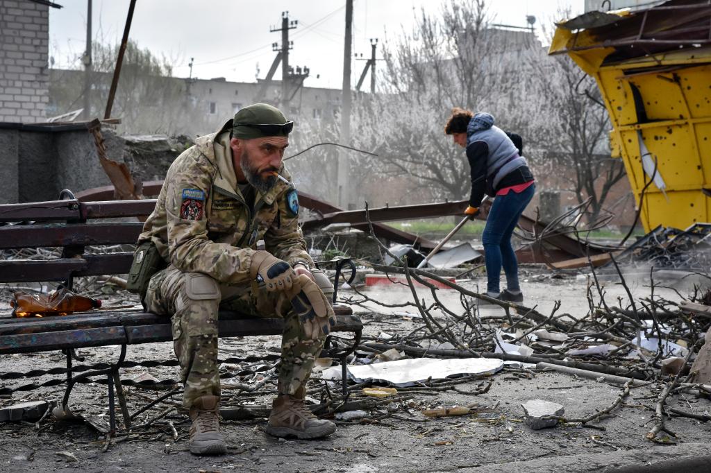 A Ukrainian serviceman wearing battle fatigues sits on a bench smoking as a local resident clears debris from a damaged building nearby.