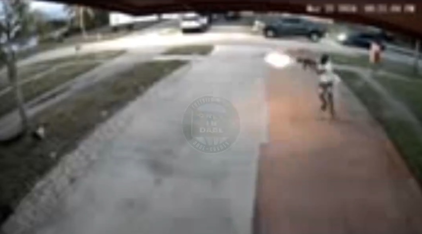 Grainy surveillance footage showing a man on a sidewalk with a muzzle flash coming out of an automatic rifle as he fires.