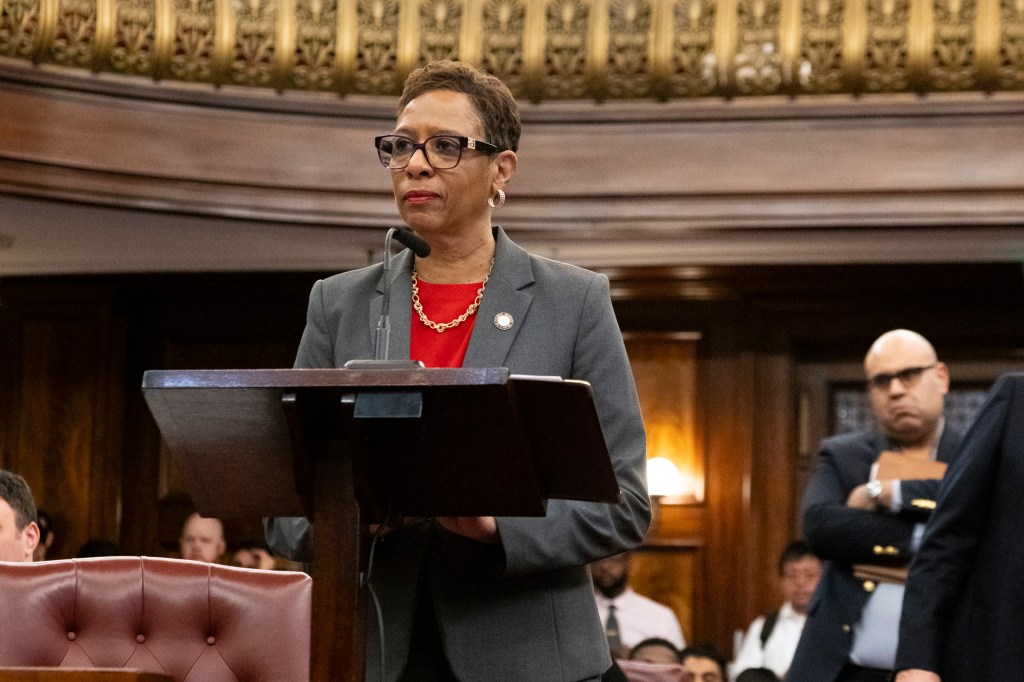 City Council Speaker Adrienne Adams standing at a lecturn wearing a grey jacket, over a red shirt and a gold necklace
