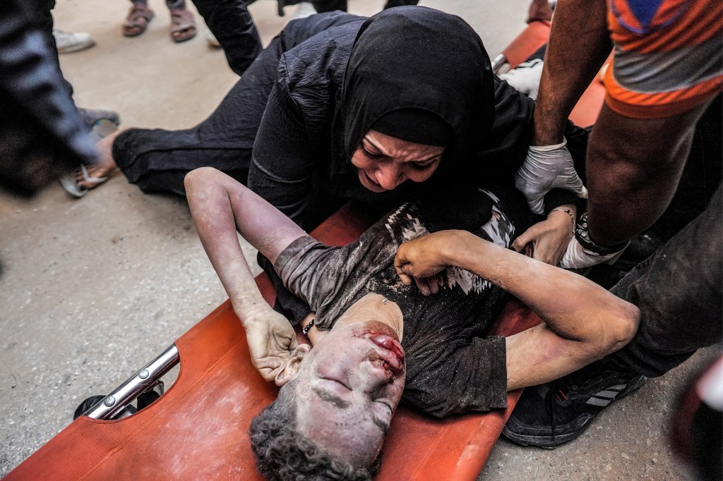 A woman in a black headscarf cries over a badly wounded or dead young child laid out on a stretcher.