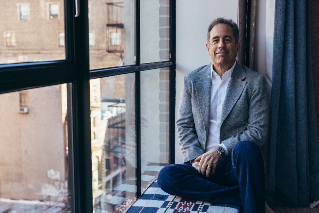 Jerry Seinfeld sitting near a window.