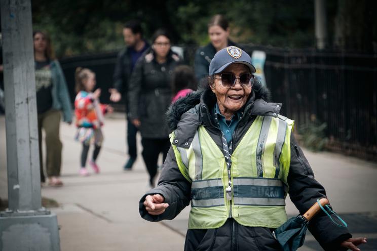 Maggie Poston on the sidewalk in sunglasses and yellow vest, families in the background.