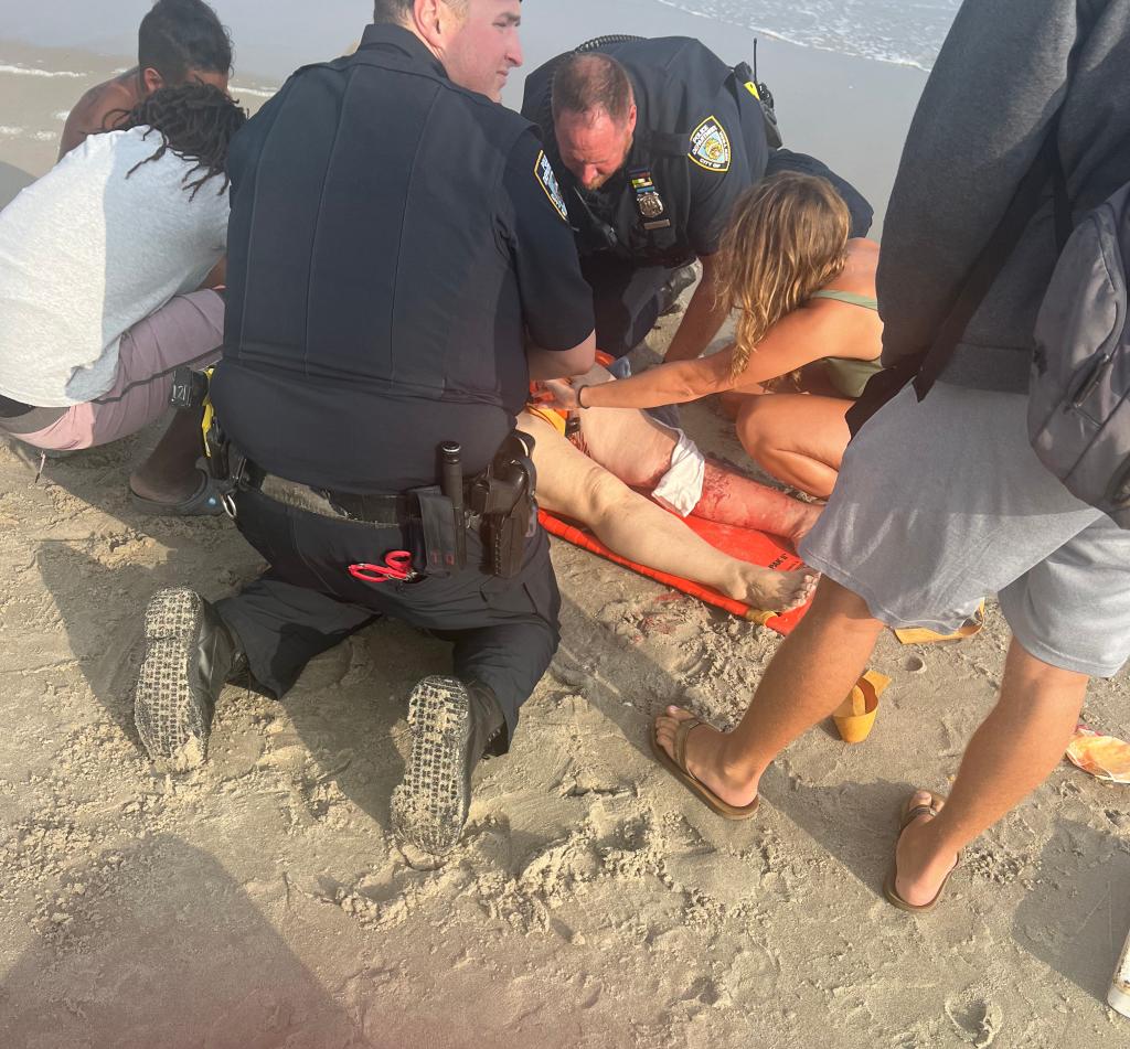police officers and beachgoers attend to a victim of a shark bite, seen here laying on an orange stretcher on the sand with her left leg bloody and bandaged