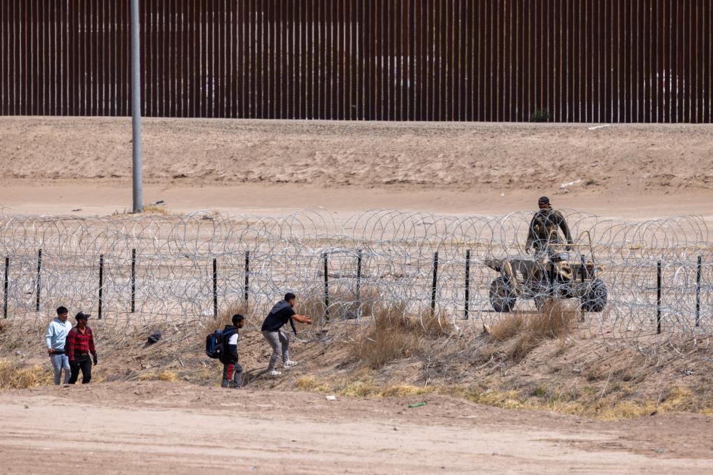 Texas National Guard pushing back a large group of migrants trying to cross the border near the Rio Grande river, with a law enforcement officer on an ATV near a concertina wire fence.