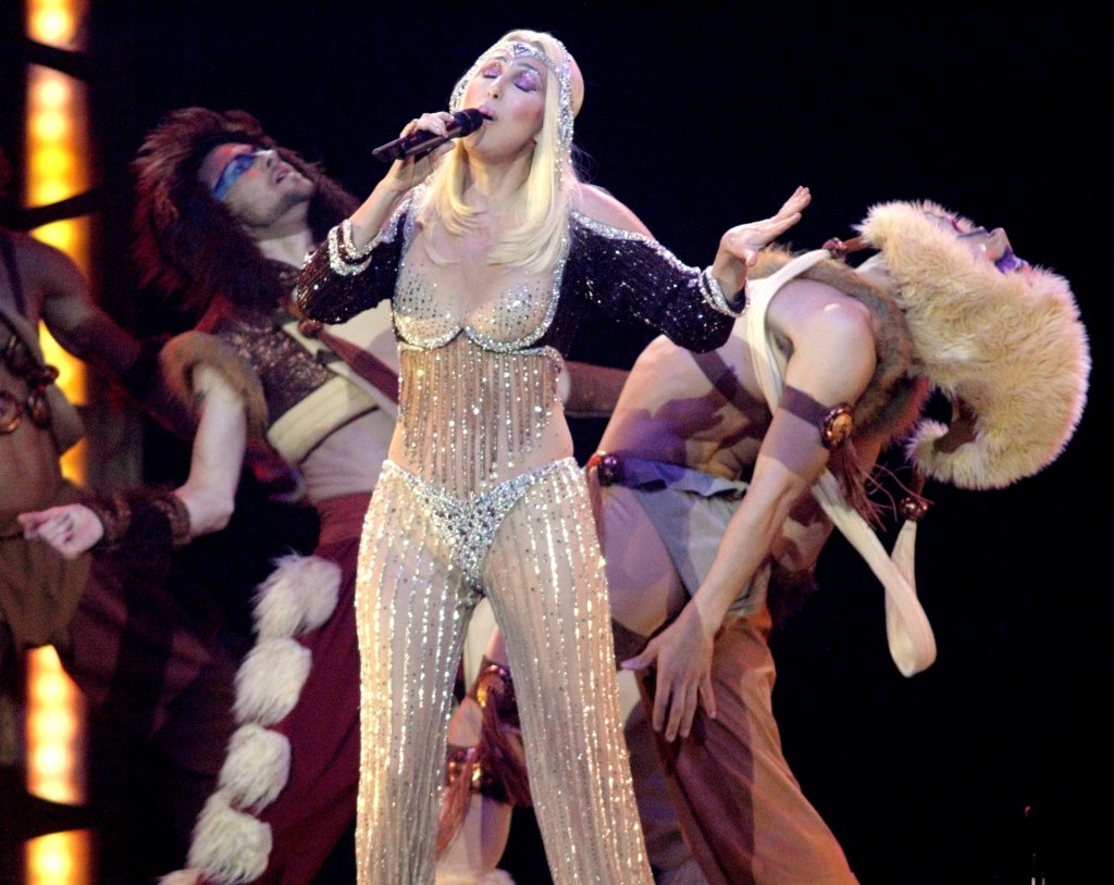 Cher wears a sheer, bewejeweled jumpsuit and sings on stage, oddly dressed dancers behind her. 