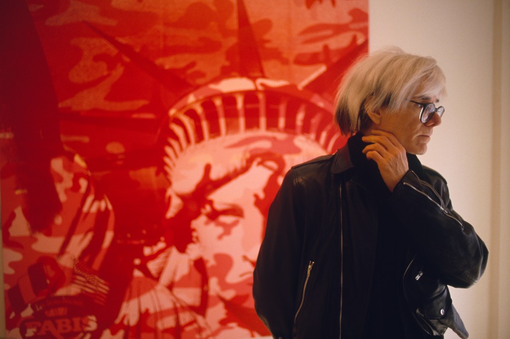 American Painter Andy Warhol, standing in front of a painting, circa 1970.