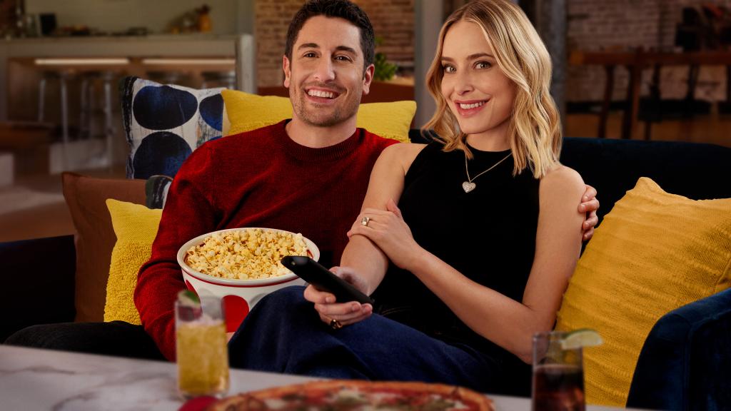 Jason Biggs and Jenny Mollen sit on a couch surrounded by yellow pillows, with biggs holding a bowl of popcorn