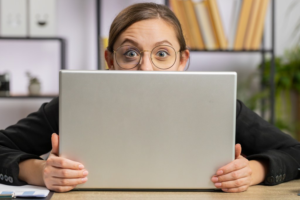 Confident business woman hiding behind a laptop computer, spying on her colleagues and looking at the camera with a cunning glance.