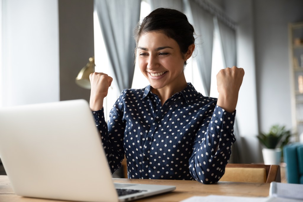 Overjoyed girl sitting at desk, reading incredible news on laptop screen, and celebrating success with clenched fists