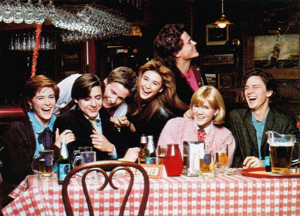 Cast of St. Elmo's Fire (1985), including Ally Sheedy, Judd Nelson, Emilio Estevez, Demi Moore, Mare Winningham, Rob Lowe, and Andrew McCarthy gathered around a table.