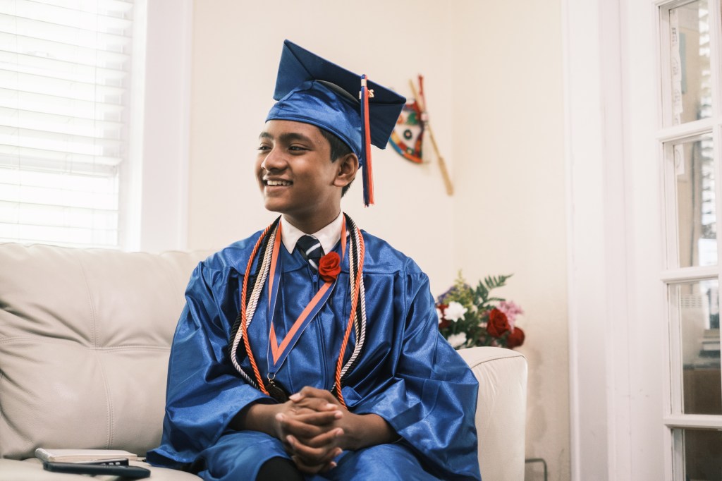 12-year-old prodigy Suborno 'Isaac' Bari in graduation cap and gown, sitting on a couch, after earning a diploma from Malverne High School