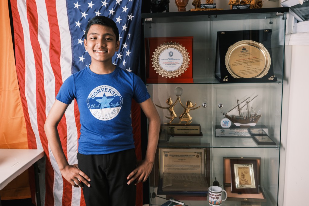 12-year-old prodigy Suborno Issac Bari standing in front of a glass display case filled with trophies, with a flag in the background.