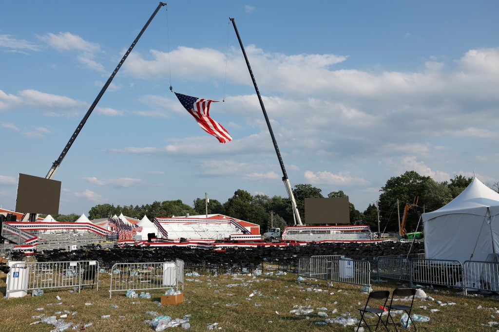 Campaign signs and discarded water bottles on the ground at a Donald Trump rally in Butler, Pennsylvania, with a flag being lifted by a crane in the background.