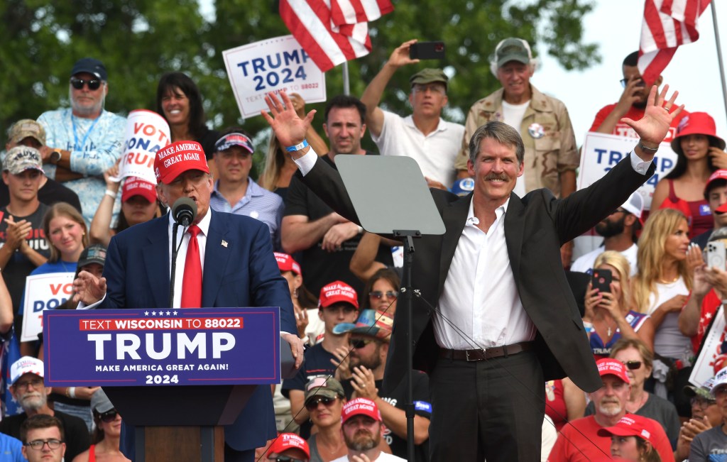 A man on the right holds his hands up in the air, next to a man on the left, who stands behind a podium and wears a "Make America Great Again" hat. The podium has a sign that says "Text Wisconsin to 88022 Trump Make America Great Again! 2024." A crowd with signs and hat smiles and waves behind the two men. 