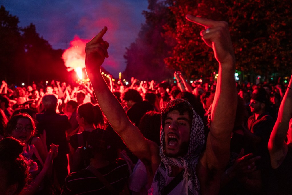 A man wearing a muslim scarf give double middle fingers as a crowd surrounding him glows red by the light of a flare.