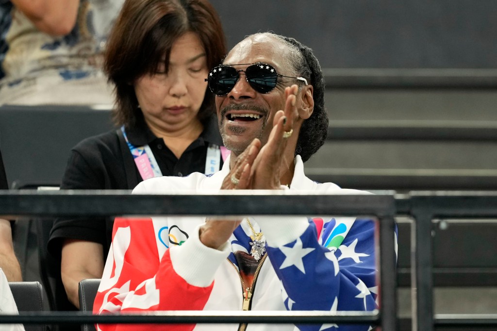 Snoop Dogg wearing sunglasses and a red and white jacket with a blue and white flag, attending the women's artistic gymnastics qualification round at the 2024 Summer Olympics in Paris.