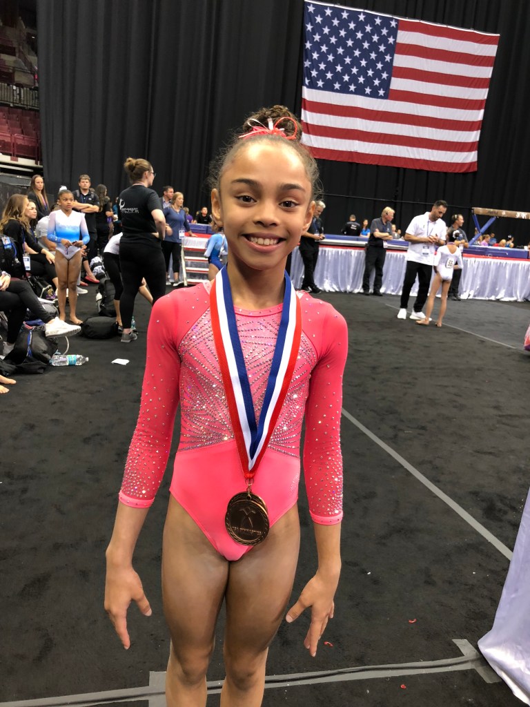 Young gymnast Hezly Rivera in pink leotard holding a medal, training at ENA Gymnastics. Photo credit to Craig Zappa.