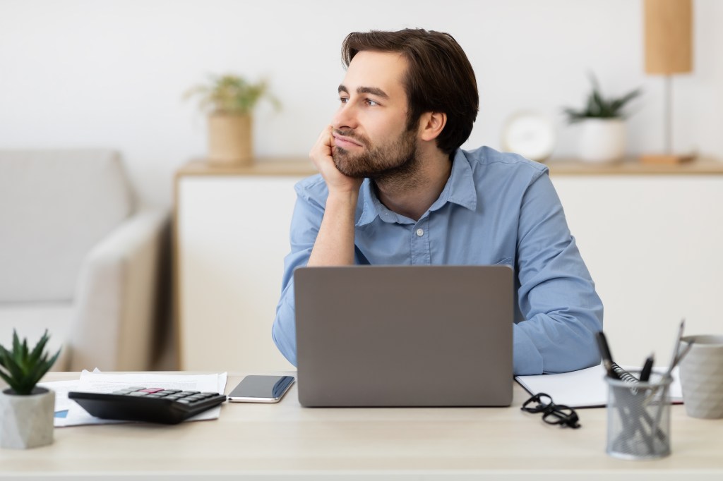 Bored businessman sitting at his desk, procrastinating and looking away from his laptop in a modern office