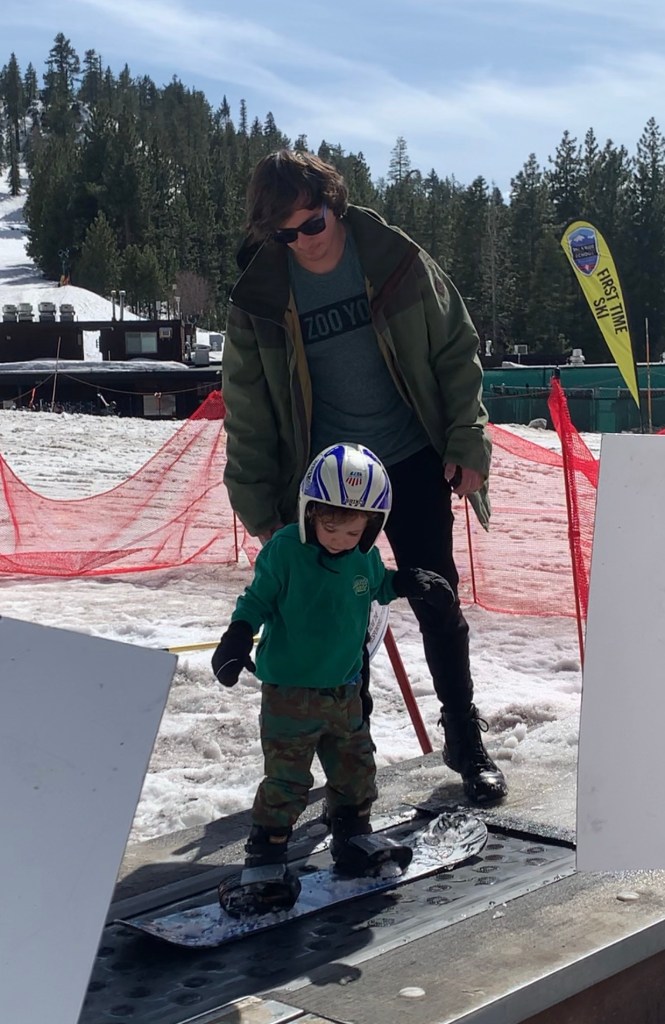 Harrison Tinsley and his 4 year old son Sawyer, father helping son stand on a snowboard.