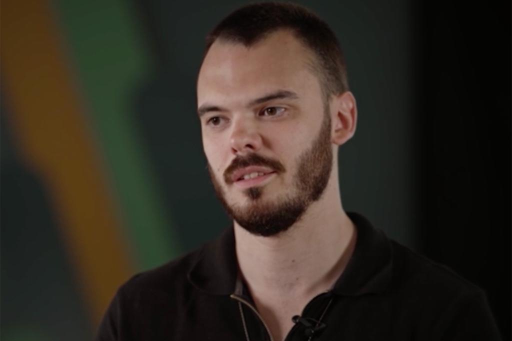 Screenshot of Andrey Kozlov on camera in interview, seen wearing black collared shirt with beard and mustache, looking off camera