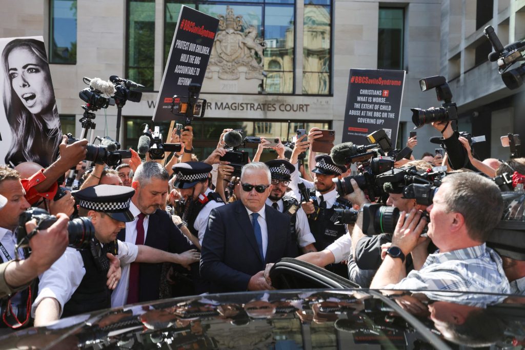 Huw Edwards, former BBC news presenter, hiding face behind sunglasses, surrounded by police officers outside Westminster Magistrates' Court, London