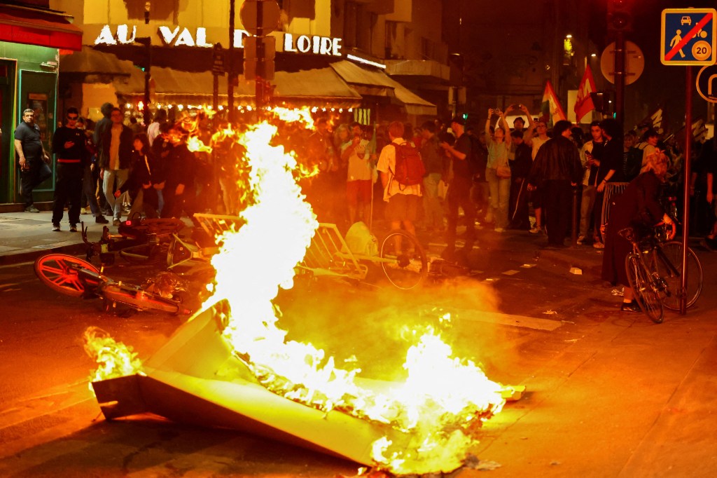 Big trash can fire in the middle of a paris street at night surrounded by onlookers.