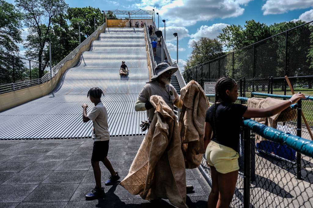 An older man wearing a wide-brimmed hat holds burlap sacks in his hands, as two young people wait at the bottom of the giant slide in Detroit, which can be seen in the background with one rider heading down. 